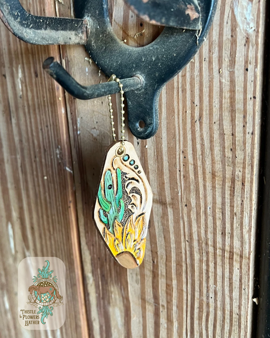 Hand tooled and painted leather keychain in vintage hotel keychain shape. Keychain has a tooled and painted green cactus and a tooled and painted yellow sunflower with a brown center. Keychain also has traditional western tooling and brown edge finish. Keychain is hanging on a stall front. Logo is in bottom right of photo.