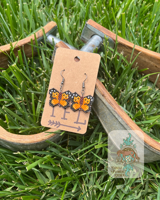 Beaded earrings that look like miniature monarch butterflies. Beads are a mix of black, a variety of oranges, white, and brown beads. Photographed on wooden and metal stirrups on grass. Logo is in bottom right of photo.