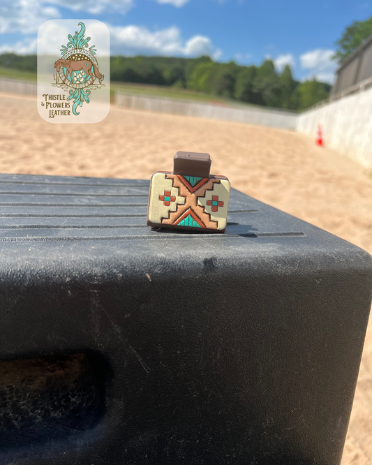 Medium brown base hairclip with hand tooled and painted leather patch. Patch is tooled with a southwestern design and the paint colors are cream, turquoise, and metallic copper. Hairclip is resting on mounting block in arena. Logo is in the top left of photo.