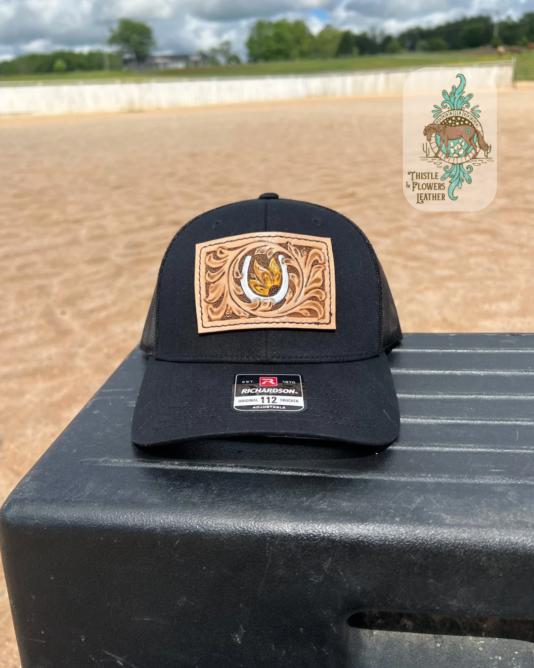 Black baseball hat with a rectangular tooled leather patch. Patch is hand tooled and painted, and features traditional western tooling, a metallic silver horseshoe, and a yellow sunflower with a brown center. Hat is on a mounting block in an arena. Logo is in top right of photo. 