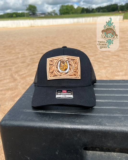 Black baseball hat with a rectangular tooled leather patch. Patch is hand tooled and painted, and features traditional western tooling, a metallic silver horseshoe, and a yellow sunflower with a brown center. Hat is on a mounting block in an arena. Logo is in top right of photo. 