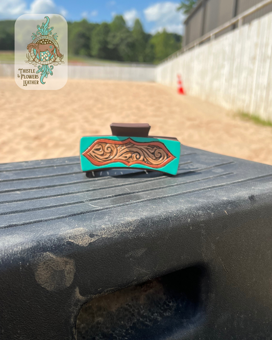 Our “Galena” hairclip sitting on a mounting block in an arena. Hairclip is hand tooled and painted. Hairclip has a peekaboo window effect with a turquoise exterior and a copper border and a hand tooled traditional western interior. Logo is in the top left of photo. 