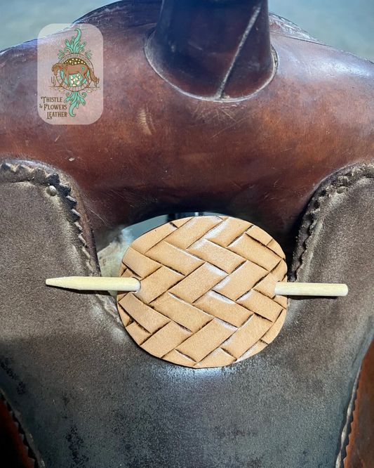 Oval hand tooled leather hairpin with light colored wooden dowel. Angled basketweave pattern is tooled in hairpin. Hair pin is sitting on western saddle. Logo is on top left corner of photo.