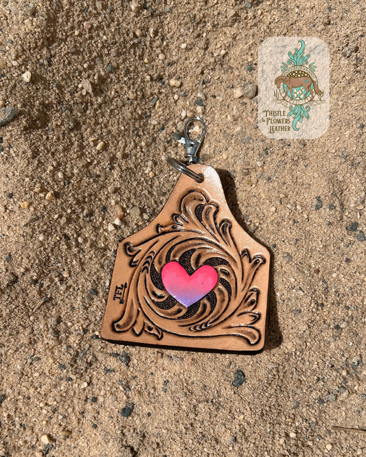Hand tooled and painted keychain with traditional western swirls around a heart. Heart is painted in an ombré pattern from hot pink to purple, top to bottom. Keychain is the size and shape of a cowtag. Keychain is in arena sand. Logo is on top right of photo.