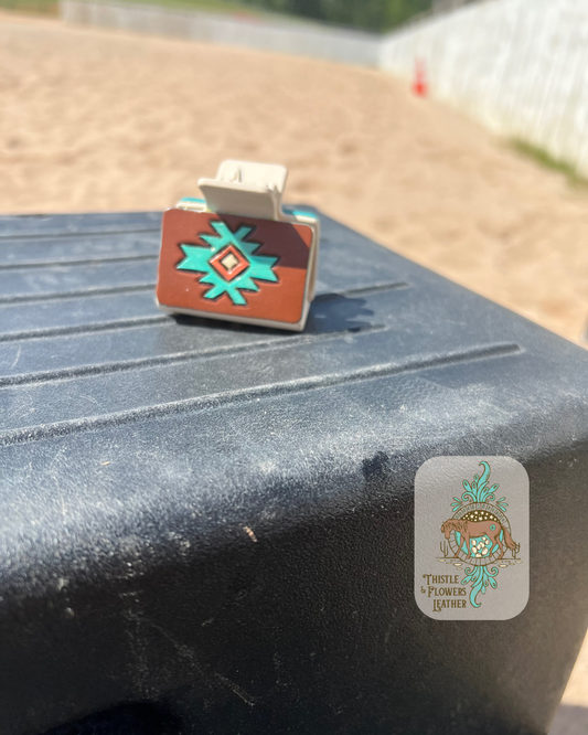 Medium cream base hairclip with hand tooled and painted leather patch. Patch is tooled with a southwestern design and the paint colors are brown, cream, turquoise, and metallic copper. Hairclip is resting on mounting block in arena. Logo is in the bottom right of photo.