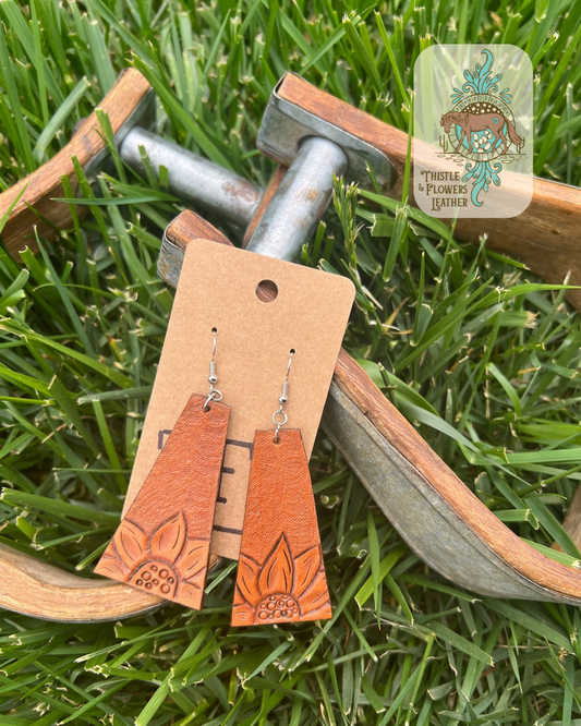 Tooled leather earrings in a long trapezoid shape with a sunflower on the bottom of each and backgrounding on the rest of  the earring. Photographed on metal and wooden stirrups on grass with logo on top right of photo.