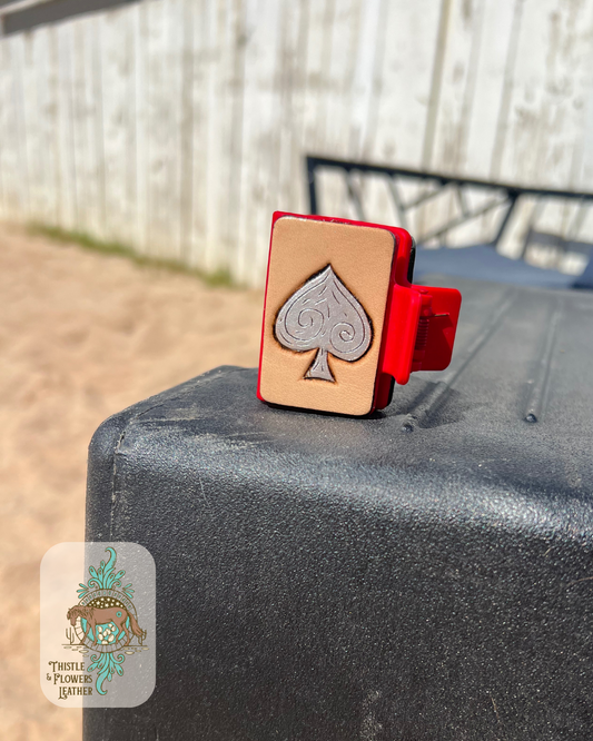 Red base Hairclip with a hand tooled and painted spade design. Spade is painted silver and has accent cuts in the leather. Hairclip is sitting on a mounting block in an arena. Logo is in bottom left of photo.