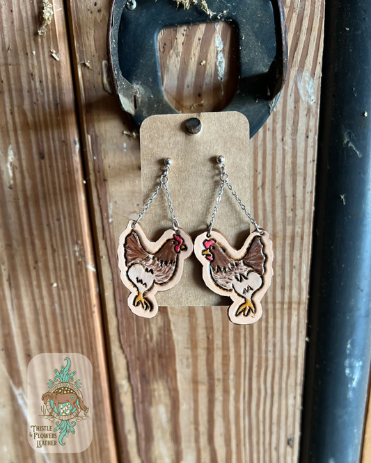 Silver colored stud earrings with 2 silver colored chains connecting to right above the chickens head and tail on each earring. Chickens are brown, red, yellow, and buff colored. Chicken earrings are hand painted and tooled. Earrings are hanging on stall front. Logo is on bottom left corner of photo.