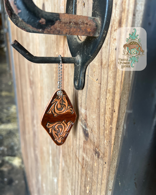 Hotel style hand tooled leather keychain. Keychain has two traditional tooling designs, one on each end. Non-tooled parts are dyed brown. Keychain is hanging on a stall front. Logo is in the top right of photo.
