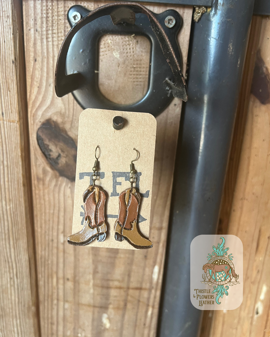 Hand tooled and painted cowboy boots shape leather earrings. Boots are a darker brown and a lighter brown color. Hardware is copper colored. Earrings are hanging on stall front. Logo is in bottom right of photo.