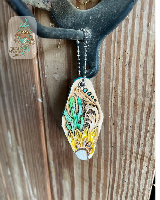 Hotel keychain shape with tooled and painted leather. Keychain has a cactus, sunflower, and traditional western tooling. Cactus is painted green and sunflower is yellow with brown center. Keychain is hanging from stall front. Logo is in the top right of photo.