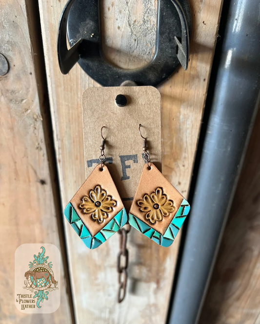 Tooled and painted leather diamond shaped earrings. Bottom edges have a tooled faux turquoise border and there is a yellow traditional western flower in the center. Earrings are hanging on stall front. Logo is the bottom left corner.