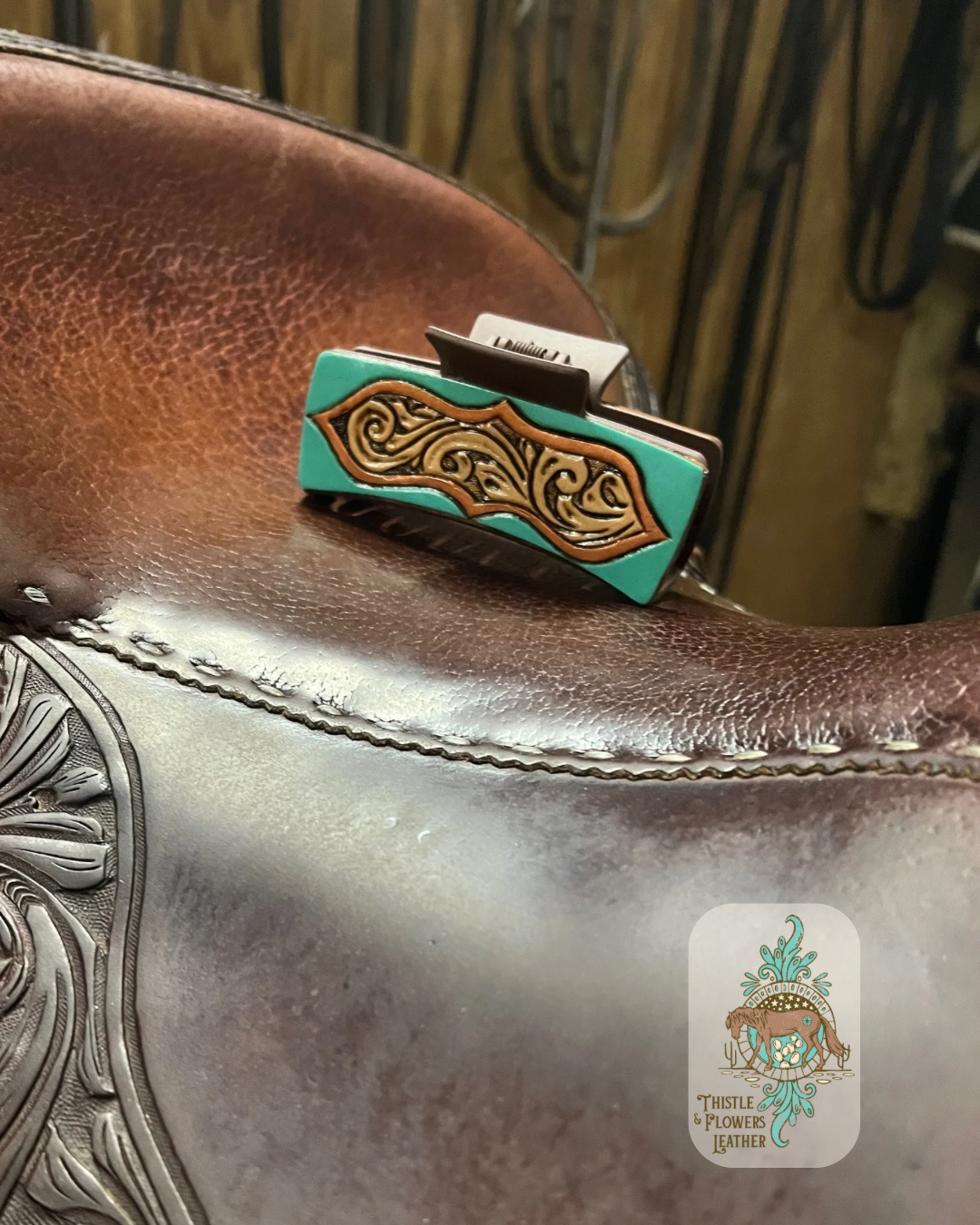 Our “Galena” hairclip sitting on a saddle seat. Hairclip is hand tooled and painted. Hairclip has a peekaboo window effect with a turquoise exterior and a copper border and a hand tooled traditional western interior. Logo is in the bottom right of photo. 