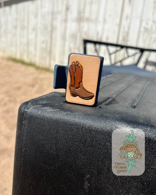 Blue base hairclip with a hand tooled and painted leather patch. Patch has a cowboy boot tooled and painted on it. Boot is painted various shades of brown. Clip is sitting on a mounting block in a riding arena.  Logo is in the bottom right corner.