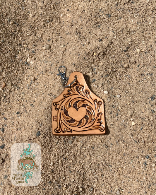 Hand tooled keychain with traditional western swirls around a heart. Keychain is the size and shape of a cowtag. Keychain is in arena sand. Logo is on bottom left of photo.