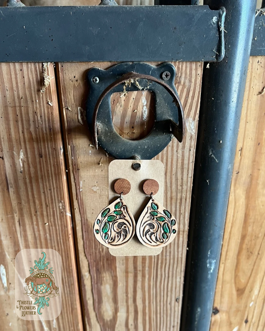 Hand tooled and painted leather earrings. Earrings feature a tooled traditional western design and a tooled and painted vine with leaves. Earrings are a teardrop shape dangling from a wooden circular stud front(actual stud-in-ear part is not wooden. Earrings are hanging from a stall front. Logo is in bottom left of photo.