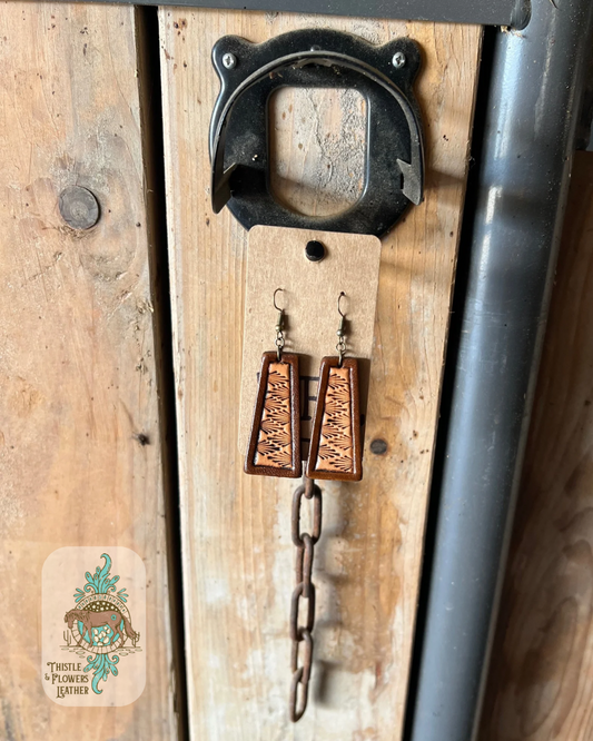 Angled bar shaped tooled leather earrings. Earrings have a brown dyed border and a western stamp pattern on the interior. Earrings are hanging on a stall front. Logo is on bottom left of photo.