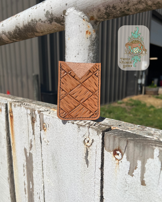 Handmade tooled leather cardholder with a large basketweave pattern. Photo shows front of cardholder  with a “v” cut for card access. Cardholder is leaning against an arena fence pole. Logo is in the top right corner of photo.