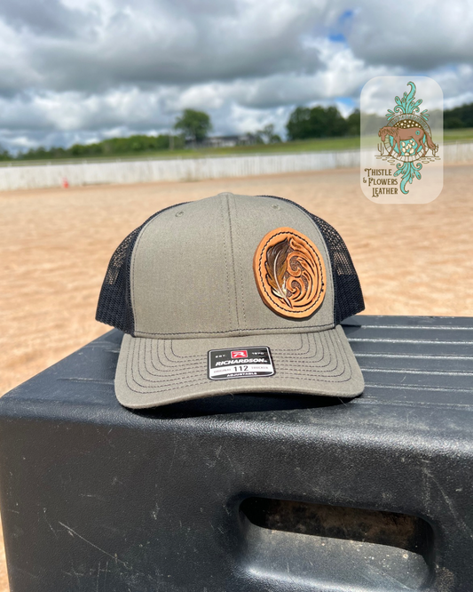 Tan baseball hat with black mesh and tooled leather circular patch. Leather patch has traditional Western tooling as well and a tooled and painted Feather. Patch is hand stitched on hat. Hat is on a mounting block in an arena. Logo is in top left of photo.