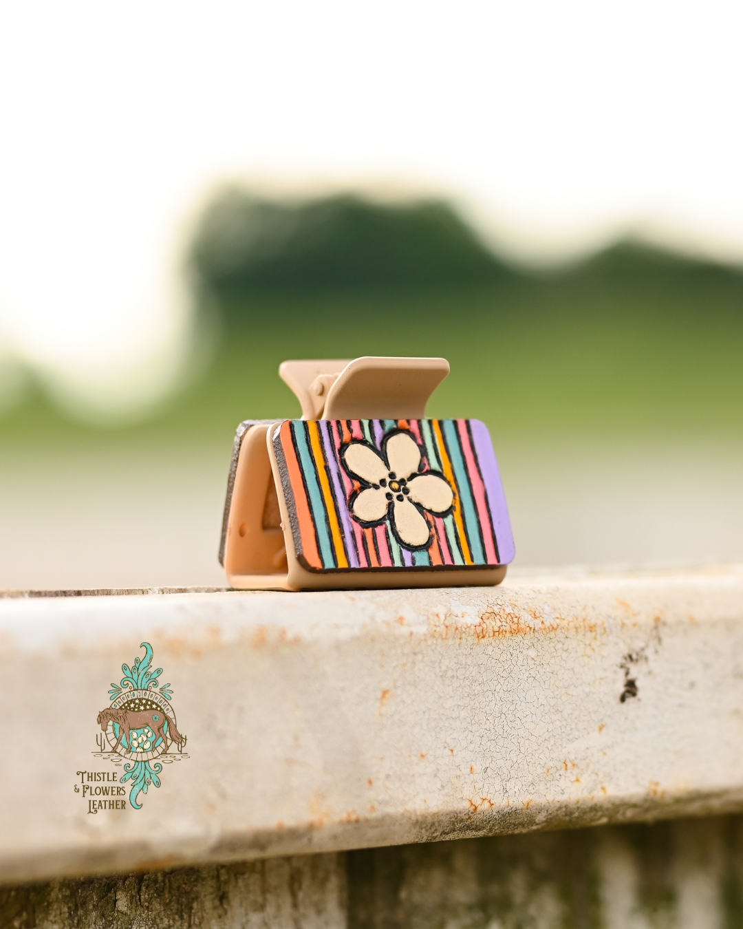 Tan medium claw clip with tooled and painted leather patches. Patches have a daisy and pastel stripes on them. Clip is resting on slightly worn metal fence.