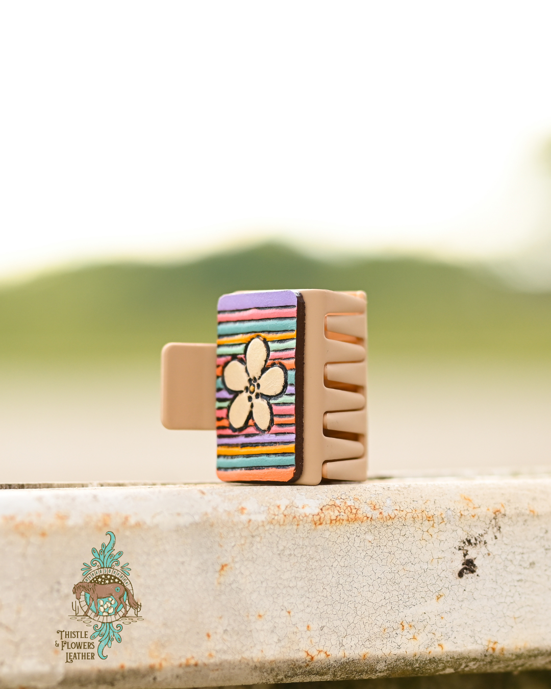 Tan medium sized rectangular claw clip with tooled and painted leather patches. Patches have a daisy and pastel colored  stripes. Teeth of hairclip are visible from this angle. Clip is resting on worn metal fence.