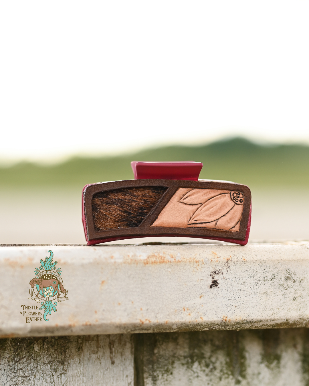 Brick red hair clip with tooled leather patches. Leather patch has a tooled flower and a brown and black cohide inlay. Clip is on a worn white fence.