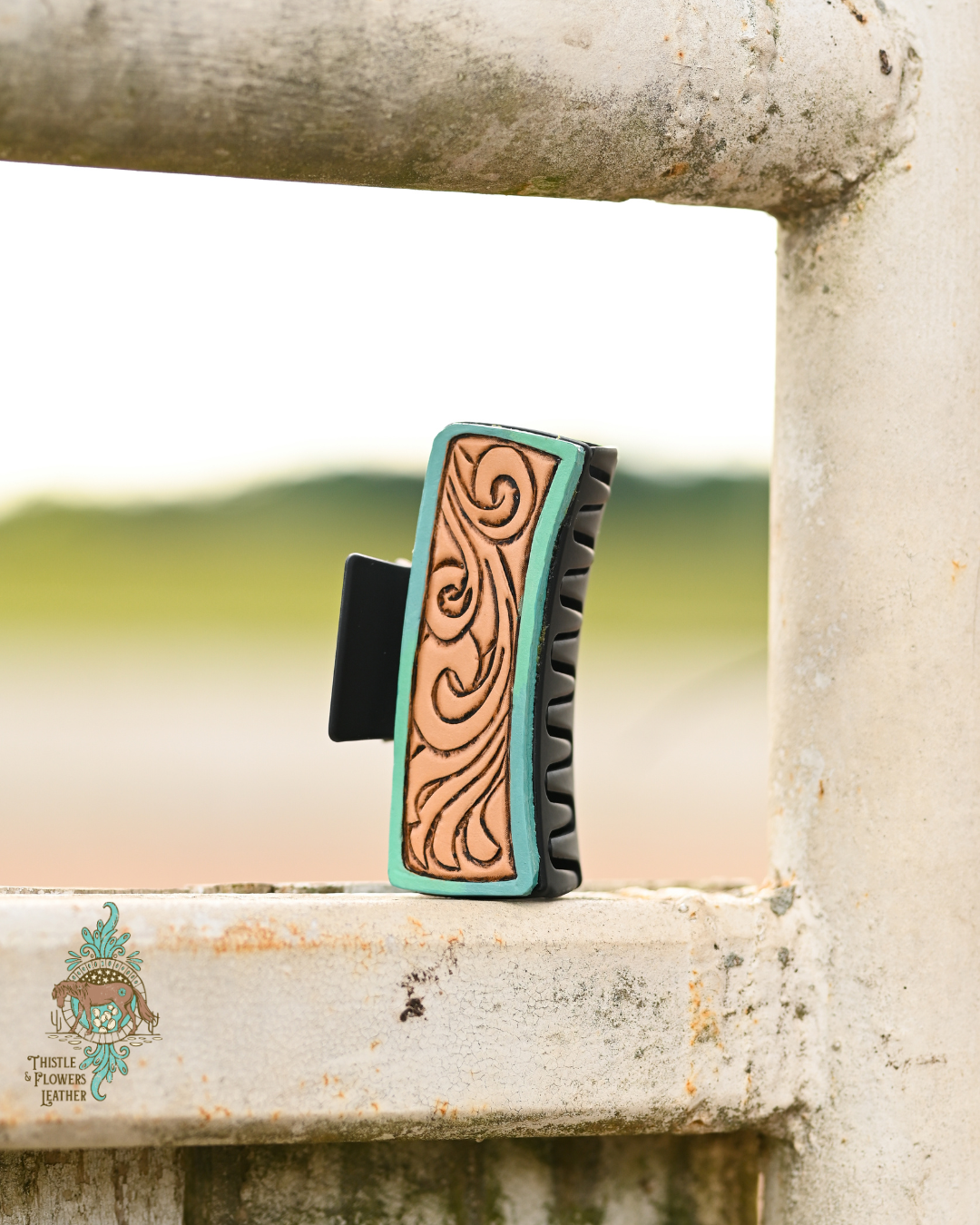 Black large claw clip with tooled and painted leather patches. Leather patches have a gradient painted from turquoise to light blue border, with traditional leatherwork scrolls flowing on the rest on the clip. Clip is on a metal worn fence.