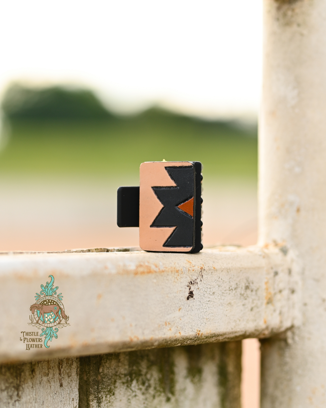 Black plastic hair clip with a tooled and painted leather patch. Patch has an Aztec design, painted black, with a rust colored 1/2 diamond in the center. Clip is sitting vertically on a worn metal fence. 