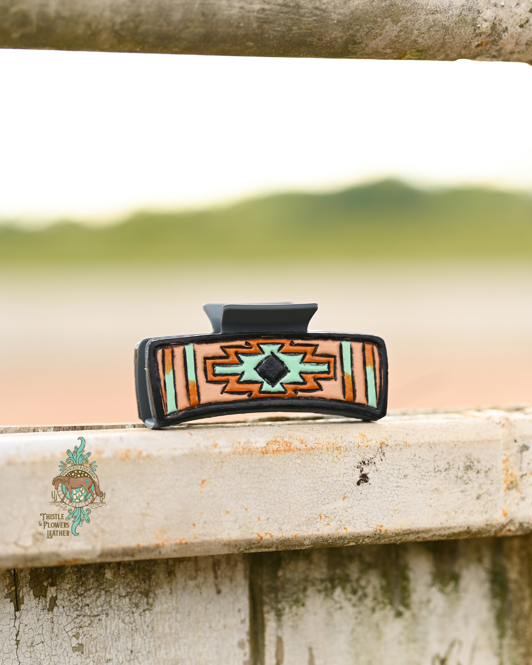 Grey large claw clip with leather patches. Tooled leather is painted in an Aztec-inspired design with colors black, mint, and a metallic copper. Clip is perched on a worn metal fence.