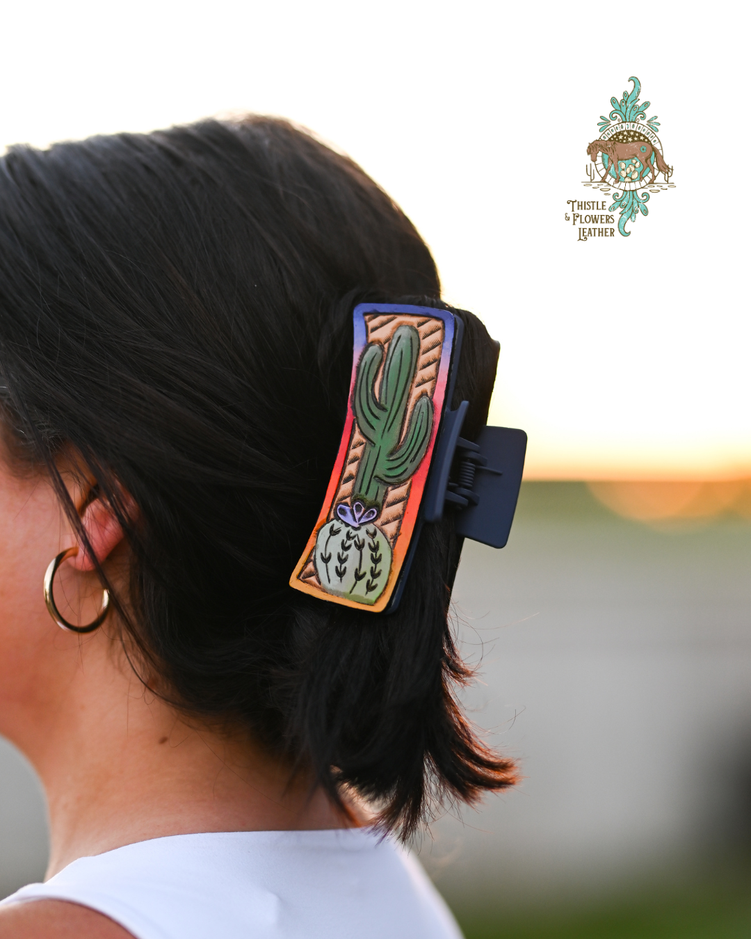 Woman with dark hair, woman has a tooled and painted leather claw clip.  Clip has a sunset gradient on border of the clip, with 2 cacti on the inside.