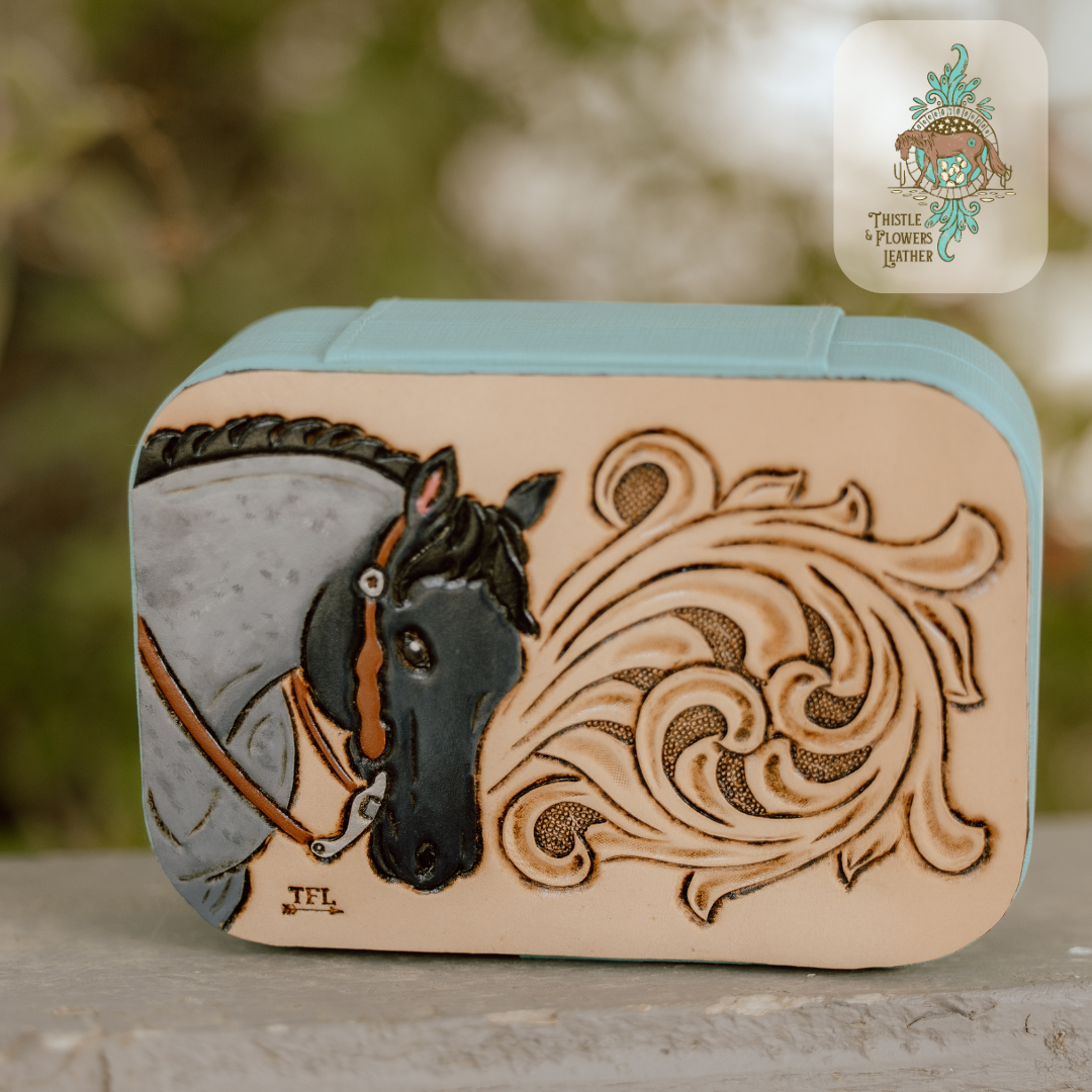turquoise travel jewelry box, medium sized with a hand tooled and painted leather patch. Patch has a tooled and painted blue roan horse with a brown headstall and reins and metallic silver bit. On other side of patch there is some traditional western tooling. Jewelry box is photographed in soft natural light in a gently out of focus background.