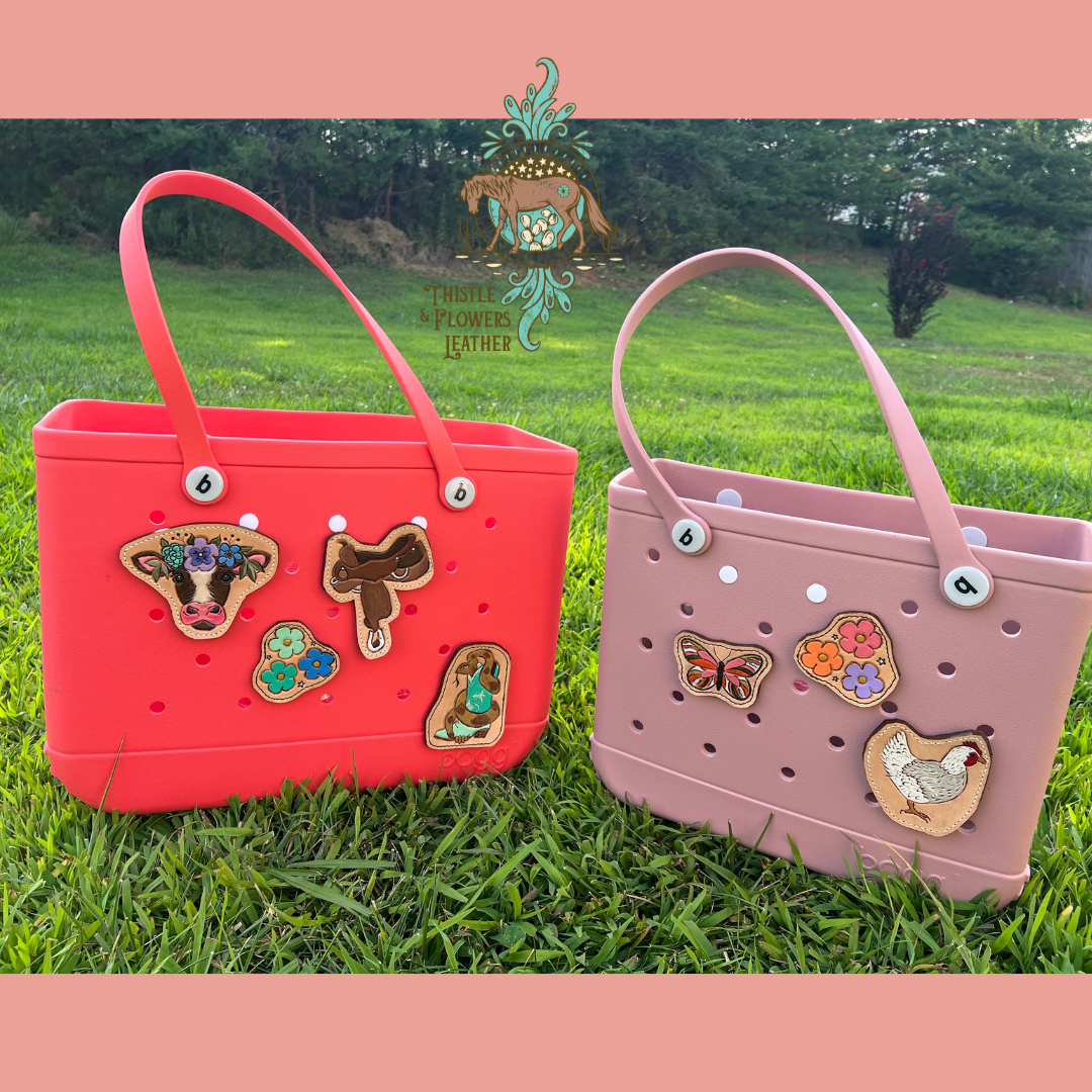 two Bogg bags in a bright green yard. Left bogg bag is hot pink, with 4 charms on it. They are a cow with a flower crown, a western saddle, blue and green flowers, and snake wrapped around a mint green boot. The bogg bag on the right is light pink and smaller, with 3 charms on it. One is a pink and orange butterfly, another is pink, purple, and orange flowers, and the final one is a white and grey chicken.