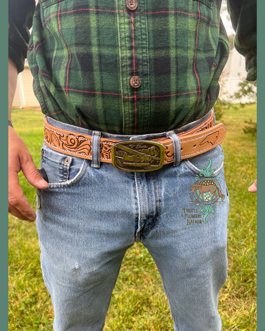 Man showing custom tooled belt from the front. Brass airplane belt buckle, tooled leather on the belt, and a tooled outline of north carolina on the tip of the belt. Man is wearing green plaid shirt and blue jeans.