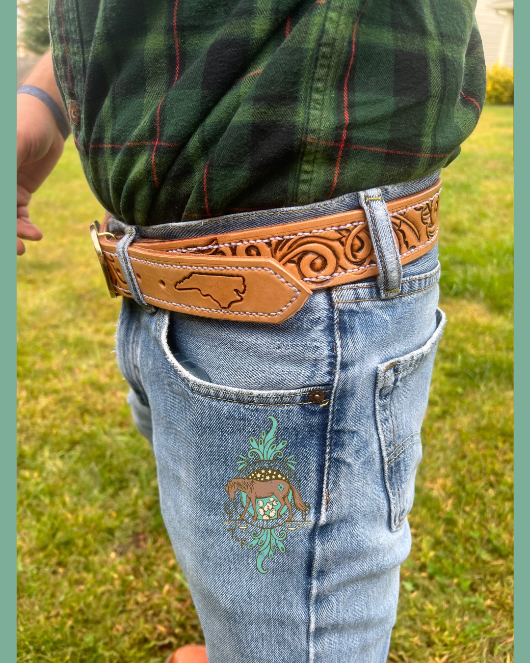 Man modeling a custom tooled belt. Man is wearing a green plaid shirt and blue jeans. Tooled vines make up the base of the belt, with the belt tip having a tooled outline of north carolina.