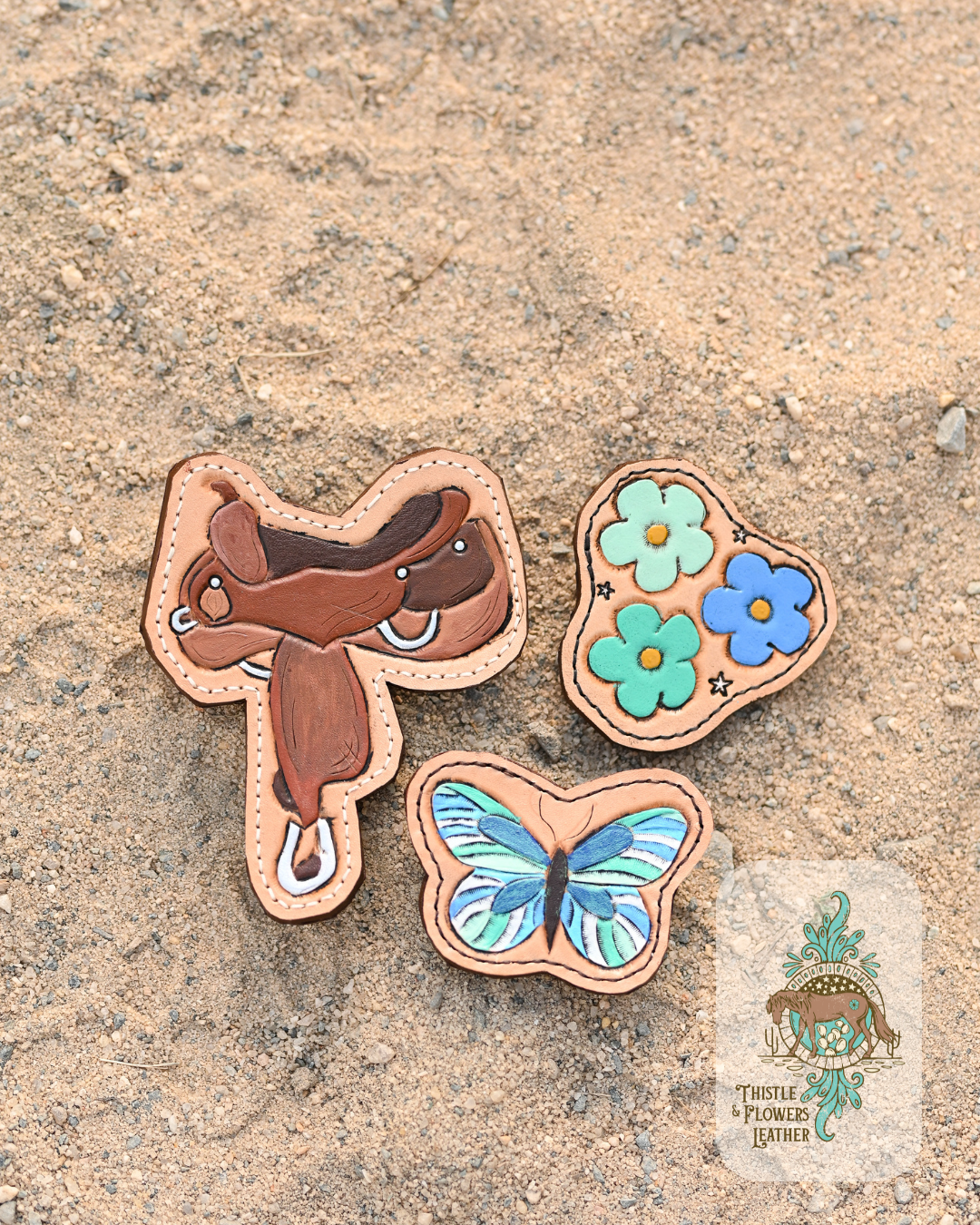 3 all weather bag leather charms on a sandy background. 1 is a tooled and painted saddle, 1 is a tooled and painted blue butterfly, and the other is 3 daisy-esque flowers in shades of mint, blue, and teal with yellow centers, and 3 small white stars.