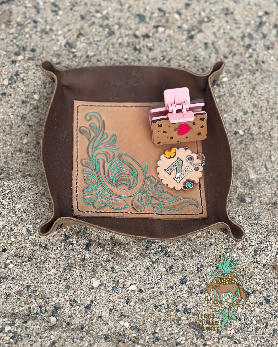 Leather valet tray with chocolate colored brown leather around exterior, and tooled veg tanned center. Tooling is of a horseshoe and floral scrolls. A hair clip and small keychain are in the tray, but not included with purchase.