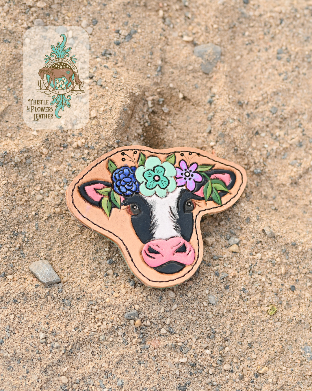 1 black and white cow tooled & painted leather all weather bag charm Cow has a flower crown with mint, blue, and purple flowers on it, as well as some greenery. Charm is laying on a sandy background.