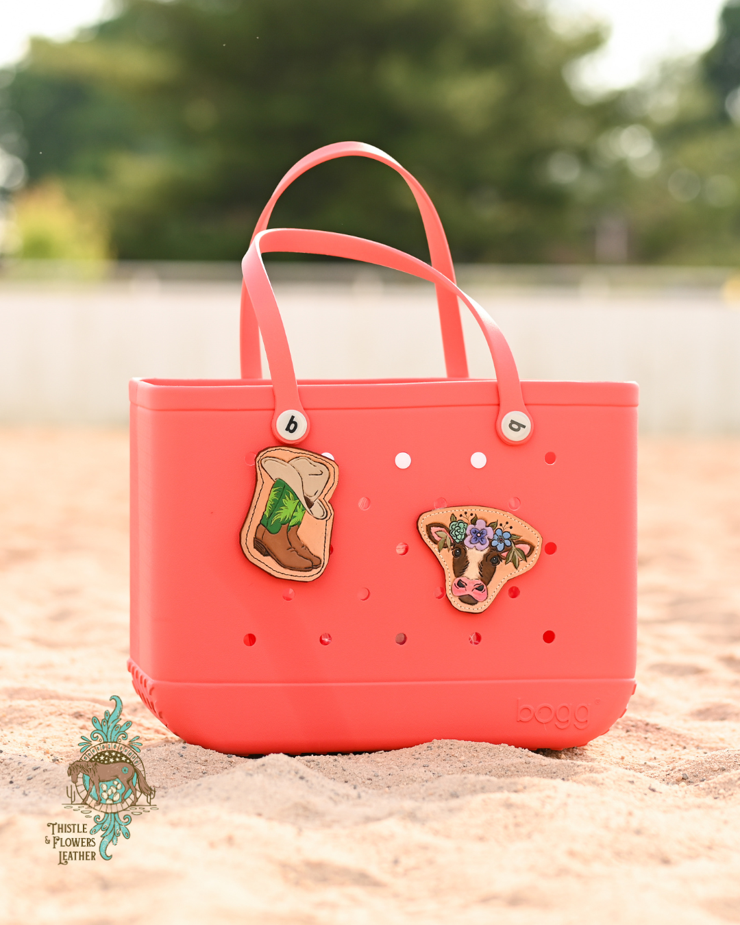 Bright coral colored large Bogg bag with 2 leather charms - 1 green cowboy boots w/a cowboy hat and 1 is a brown and white cow with a flower crown. Bag is in a sandy arena.