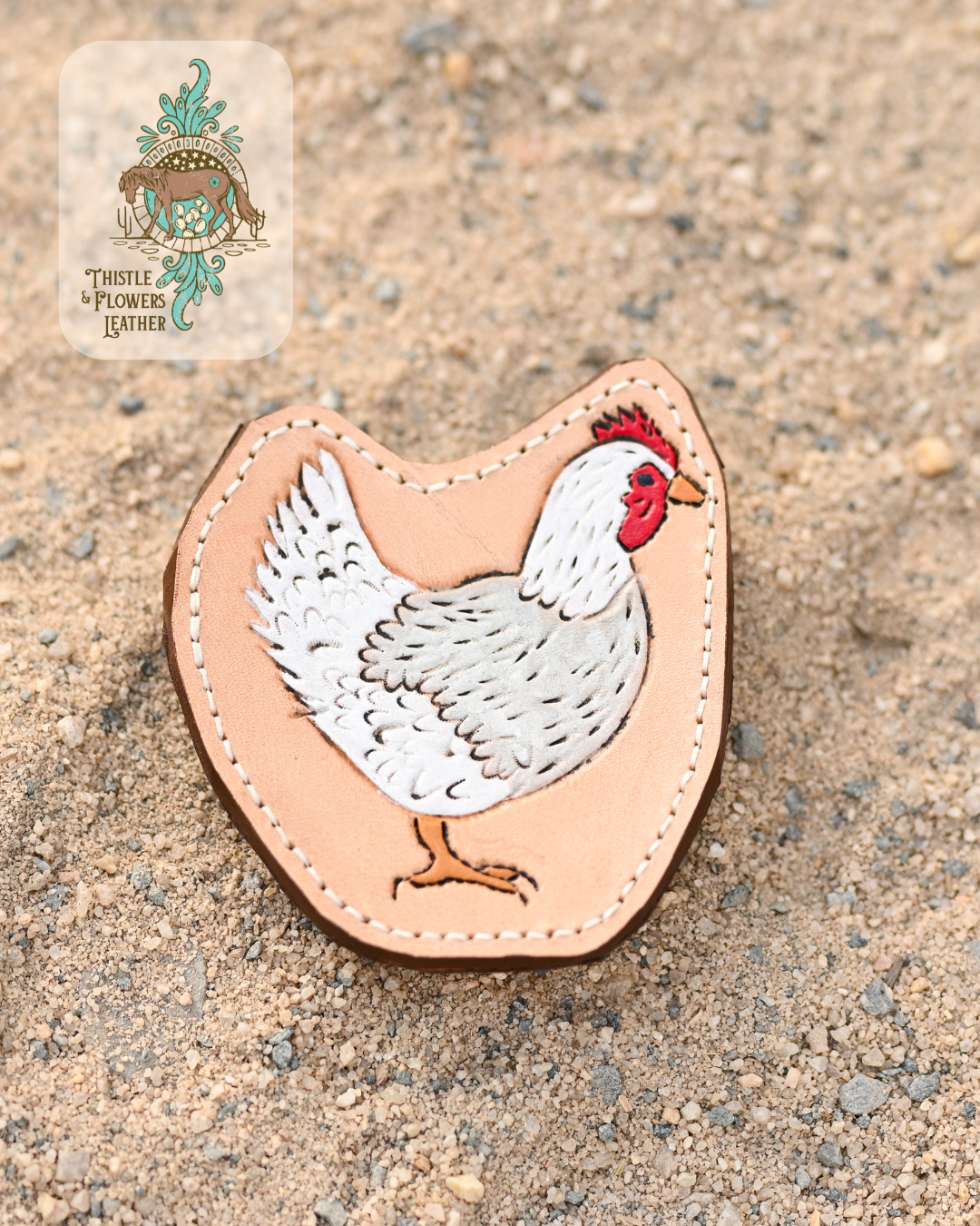 Tooled leather all weather bag charm with a chicken tooled on it. Chicken's head and tail are white, with the main body being a light grey. Chicken also has red comb on top of head and around eye/beak. Charm is on a sandy background.