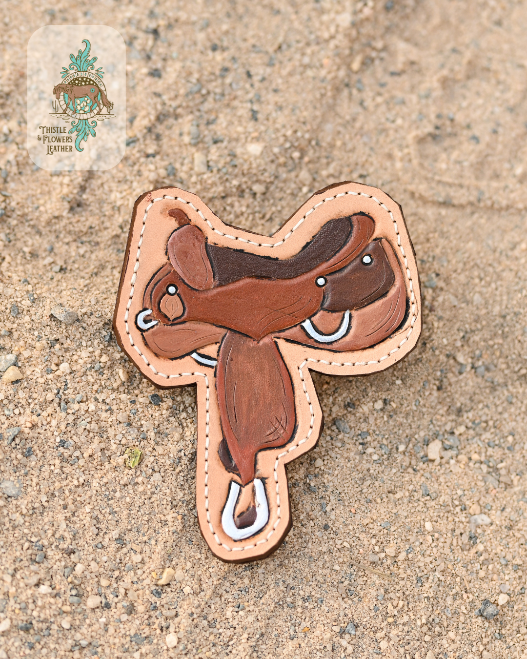 Tooled leather all weather bag charm in the shape of a western reining saddle. Hardware points on saddle are painted with a silver metallic. Saddle is painted various shades of dark brown.  Charm is on a sandy background.