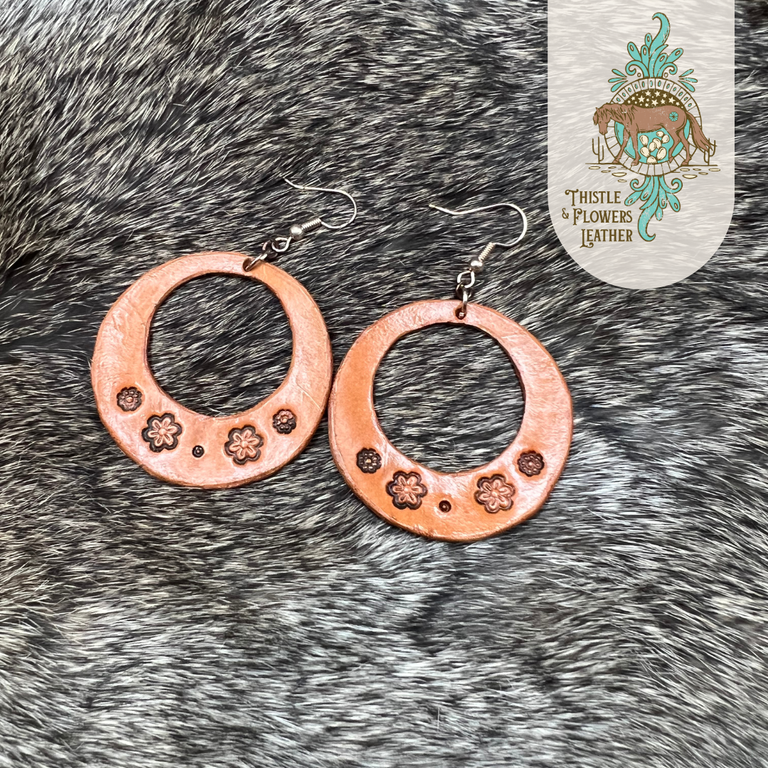 Tooled leather earrings in a circular shape with a circle cut out of the center (cutout is not perfectly aligned). Tooled leather flowers are on the bottom of the circle. Earrings are nickel-free silver colored hooks and jump rings. Earrings are laying on rabbit fur.