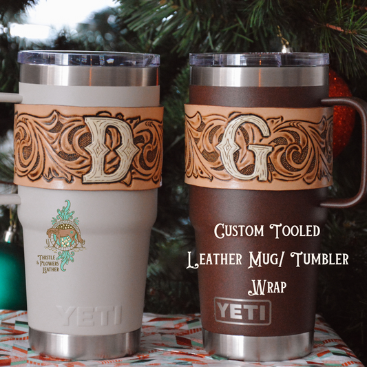 2 insulated tumblers with leather wraps with initials &quot;D&quot; on one and &quot;G&quot; on the other. Tumblers are sitting on a wrapped gift, with Christmas tree branches in the background.