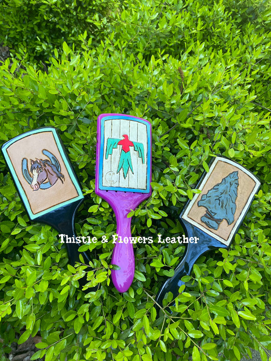 3 full size hairbrushes with tooled leather patches on a bright green brish. the left brush has a black brush with a tooled leather patch that has a  bay horse in a silver horseshoe tooled and painted in the center, and a mint border around the patch. The middle brush is purple with a tooled patch with a mint and hot pink thunderbird, white background, and a blue ombre border. the right brush is black with a tooled leather patch. Leather patch has an arrowhead painted and tooled on it, with a white border.