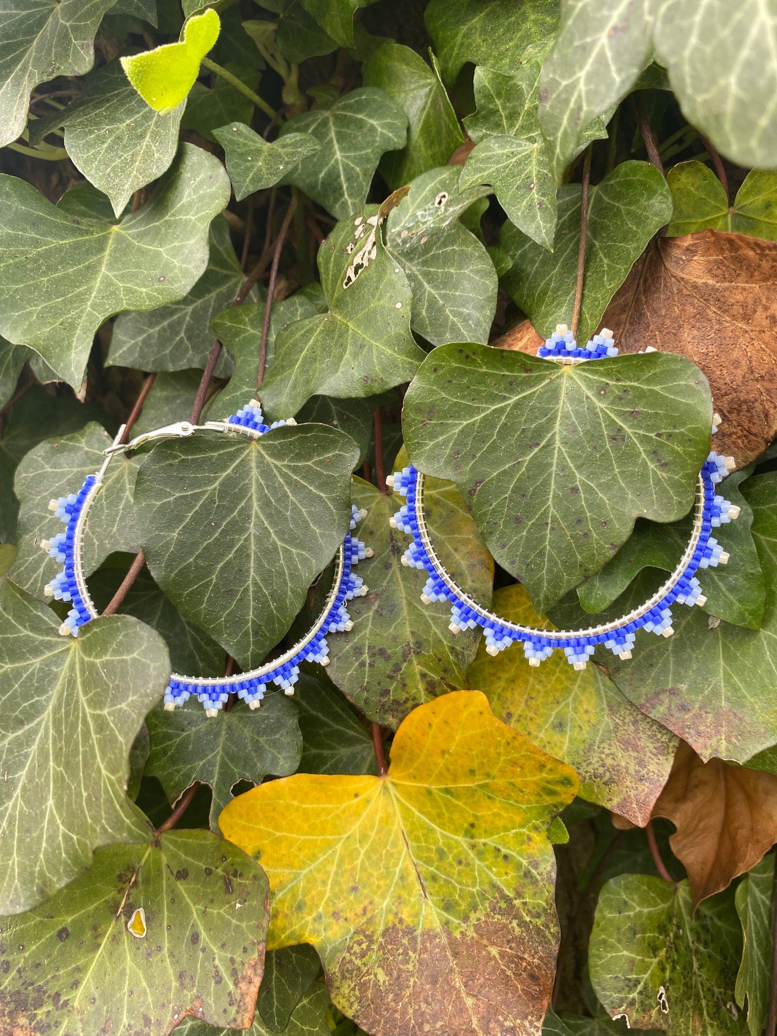 Beaded hoop earrings with a sliver colored base. With a dark blue base, the earrings have small trianglular shapes coming off the base. The tips of the triangles are white, and then lighter blue is in between the white and the dark blue. Earrings are on English Ivy.