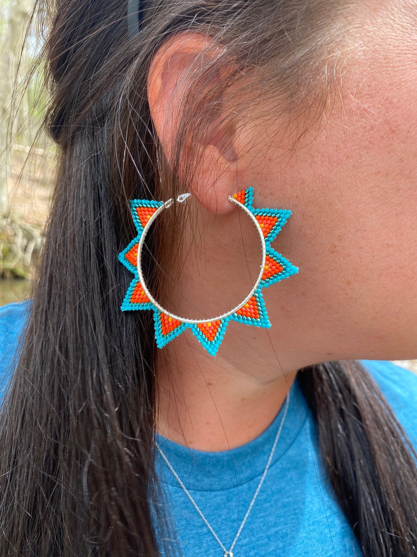 Model with brown hair is wearing hoop earrings with a silver colored base, and trianglular beading coming off the hoop base. Turquoise, metallic turquoise, dark orange, orange, and yellow beads are on the earring. The triangles are edged in tuqoise, and with am ombre effect fade from deep orange to a single yellow bead in the center of the triangle.